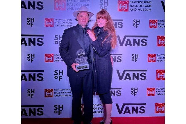 Red carpet photo of a man in a dark suit and white fedora hat, standing with his wife, who has long red hair and is wearing a long black coat and black capri pants. He is holding a small trophy shaped like half of a skateboard that reads "Skateboarding Hall of Fame" in front of a white backdrop with the Vans company logo on it.